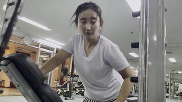 A woman wearing a white top and short skirt, bent over a piece of exercise equipment. She appears to be working out in a fitness center. The gym has other exercise equipment and mirrors. video