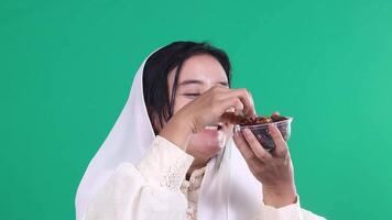 A woman holds a date in one hand and a container of dates in the other, while wearing a white headscarf and dress. isolated on a green background or green screen. video