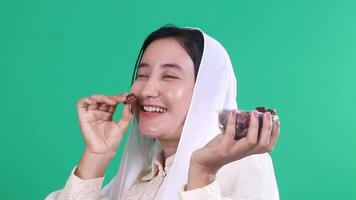 A woman wearing a white headscarf is holding a container of dates, picking one out to eat. isolated on a green background or green screen video