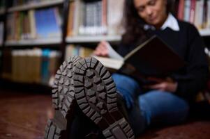 Person reading a book seated in a library with visible boot soles photo