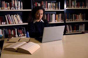 Woman studies using laptop surrounded by books in library. This serene setting reflects focused learning and academic effort, portraying the essence of knowledge acquisition, education in quiet space photo