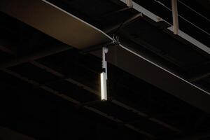 Illuminated Industrial Elegance A sleek, elongated ceiling light pierces the shadows of a dark metal web, casting a warm glow in an urban oasis. photo