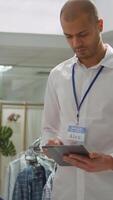 Vertical Middle eastern clothing store employee verifying merchandise stock on device, preparing the new collection on display for customers. Ensuring a great luxurious shopping experience. Camera B. video