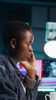 Vertical African american woman working from home, using laptop, looking at statistic charts. Remote worker in personal apartment office crosschecking important info between clipboard and notebook, camera B video