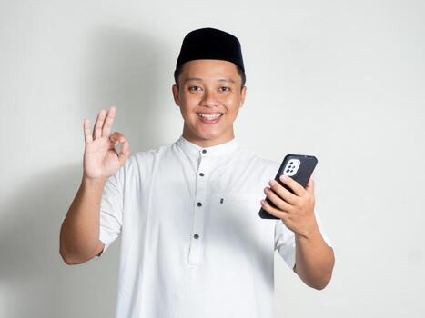 Muslim Asian man holding a phone and makes an okay hand gesture photo