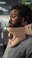 Vertical Hurt employee with a cervical collar working on a new business project, struggling to sit at the desk with his neck fracture. African american company associate tries to solve tasks. Camera A. video