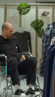 Vertical Middle eastern client in a wheelchair examining new clothes on racks at the retail store, consumerism and style choice. Young man with a chronic disability shopping for trendy items. Camera A. video