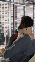 Vertical Hurt employee with a cervical collar working on a new business project, struggling to sit at the desk with his neck fracture. African american company associate tries to solve tasks. Camera B. video
