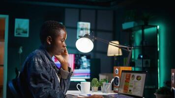 African american woman working from home, using laptop, looking at statistic charts. Remote worker in personal apartment office crosschecking important info between clipboard and notebook, camera B video