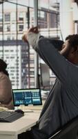 Vertical Black male CEO dealing with back pain and stretching his muscles at the desk, sitting with an incorrect posture and trying to fix it. General manager working with data analytics. Camera B. video