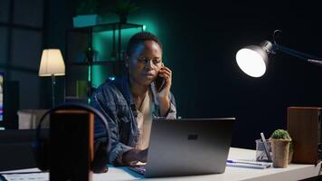 Woman receiving phone call from friend while working from home on laptop in personal office. Freelancer speaking with mate online using cellphone while solving tasks, camera B video