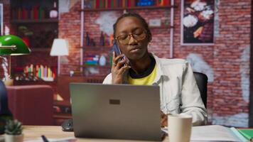 Woman in home office sitting at desk in front of laptop, answering phone call. Remote employee using notebook to do job online, receiving telephone call on smartphone, camera B video