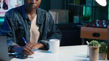 Jolly freelancer noting down ideas on paper using pen while teleworking. Upbeat remote employee working at home office desk, reading information on laptop and writing in notebook, camera A video