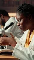 Vertical Dedicated female student working with a microscope to prepare for microbiology lessons in a library, focusing on microorganisms and chemical processes. Notes for her academic project. Camera B. video