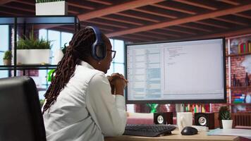 Jolly engineer in home office working on computer, dancing at desk in celebration after successfully executing code. IT worker wearing headphones happy after finishing compiling code, camera A video