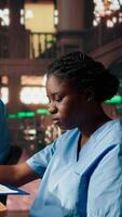Vertical African American nurse in training reviews anatomy case files in a vintage library. Med school student in scrubs studying from database books and research papers in the campus. Camera A. video