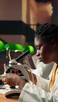 vertical dedicado hembra estudiante trabajando con un microscopio a preparar para microbiología lecciones en un biblioteca, enfoque en microorganismos y químico procesos. notas para su académico proyecto. cámara una. video