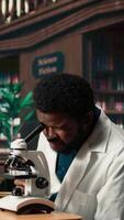 Vertical Male student researching viruses during a microscope analysis and academic reading in a classic library. Med school pupil reflects dedication to biology and exam preparations. Camera A. video