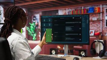 Woman using isolated screen smartphone and AI LLM chatbot on computer screen in home office. Person holding chroma key phone uses artificial intelligence chatbot on monitor, camera A video