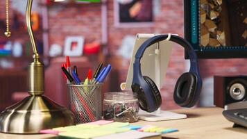 Close up of headphones on home office desk next to PC screen, stationary items, surrounded by plants. Audio device headset next to computer display in loft with shelves filled with books in background video