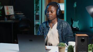 Remote worker with wireless headphones working from living room office, sending emails. Freelancer doing internet research using laptop while listening to music to make time pass faster, camera A video