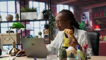 Busy freelancer in home office eating snack on the go while typing on laptop keyboard. Woman enjoying croissant while using electronic device to solve job tasks online, camera B video