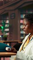Vertical Black woman student in med school using an oximeter and reading about the practice, studying with academic database copy books at the library. Scholar does her medical research. Camera B. video
