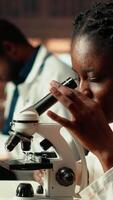 verticale femelle science étudiant examine cellules en dessous de le microscope pour sa scientifique recherche dans une Campus bibliothèque, prend Remarques à propos chimique processus et réactions pour sa thèse. caméra b. video