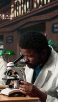 vertical africano americano aluno estudos microbiologia dentro uma campus biblioteca, examinando células e microorganismos debaixo a microscópio. masculino aluna prepara para dele biotecnologia tese. Câmera uma. video