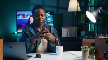 African american woman relaxing in apartment, texting friends on cellphone, enjoying leisure time. Smiling remote worker taking break to talk with mates on internet using phone, camera A video