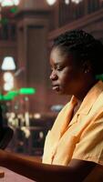 Vertical Focused african american student sits in a quiet library reading papers and class notes, studying under the warm glow of a green lamp. Woman surrounded by books and learning tools. Camera B. video