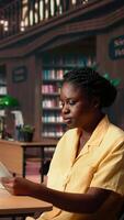 Vertical Focused african american student sits in a quiet library reading papers and class notes, studying under the warm glow of a green lamp. Woman surrounded by books and learning tools. Camera A. video
