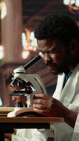 Vertical African American pupil studies microbiology in a campus library, examining cells and microorganisms under the microscope. Male student prepares for his biotechnology thesis. Camera B. video