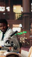Vertical Female college student focuses on medical science by using a microscope, examining the chemical processes of microorganisms. Woman studying for her academic thesis paper. Camera A. video