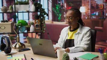 Student at home drinking coffee and browsing internet on notebook, researching information online. Woman enjoying hot beverage while reading internet articles on laptop for school tasks, camera A video