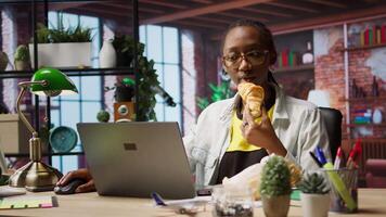 Busy freelancer in home office eating snack on the go while typing on laptop keyboard. Woman enjoying croissant while using electronic device to solve job tasks online, camera A video