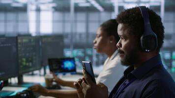 Worker in data center wearing headphones taking break from implementing AI systems, using smartphone. Focus on artificial intelligence server farm employee using mobile phone, camera A video