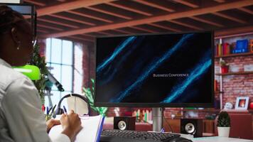 Remote worker at home watching business conference on PC monitor, looking over statistics on documents. Freelancer in apartment looking at presentation and checking paperwork files, camera A video
