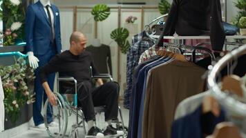 Middle eastern client in a wheelchair examining new clothes on racks at the retail store, consumerism and style choice. Young man with a chronic disability shopping for trendy items. Camera A. video
