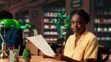 Dedicated black student reviewing papers in a traditional library setting, focusing on educational goals and study time within the campus. College girl studies and does research. Camera B. video