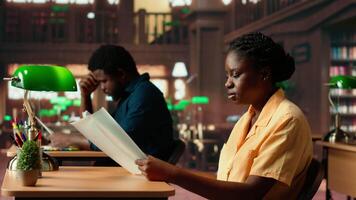 Focused african american student sits in a quiet library reading papers and class notes, studying under the warm glow of a green lamp. Woman surrounded by books and learning tools. Camera B. video