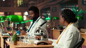 Black students team researching viruses and microbiology methods with a microscope, exchanging ideas and knowledge in the library. Takes notes and consults academic databases. Camera B. video