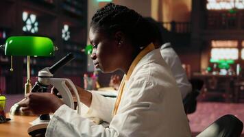 Black university student examining cells and organisms under microscope lenses, researching and studying for medical science exams. Reading about biotechnology in the library. Camera A. video