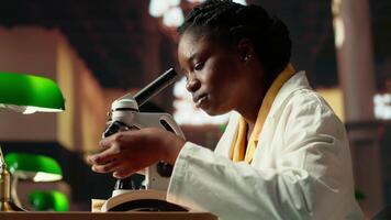 Black university student examining cells and organisms under microscope lenses, researching and studying for medical science exams. Reading about biotechnology in the library. Camera B. video
