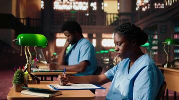 African American nurse in training reviews anatomy case files in a vintage library. Med school student in scrubs studying from database books and research papers in the campus. Camera A. video