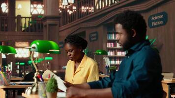African american students learning a foreign language together at the university library, consulting books from the academic database. Class colleagues helping each other study for exams. Camera B. video