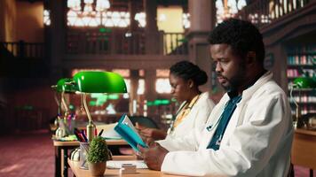 Medical student reading a book and reviewing notes from the library database, focusing on medical research. Young man reflects dedication to healthcare career and academic goals. Camera A. video