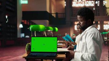African American med student reading a healthcare book next to copy space, focusing on medical practice methods under green lamps in a classic library. Commitment to education. Camera A. video