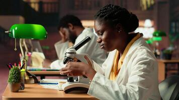 Dedicated female student working with a microscope to prepare for microbiology lessons in a library, focusing on microorganisms and chemical processes. Notes for her academic project. Camera B. video