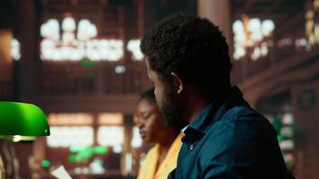 Black college students comparing their class notes at the university library, scholars team studying and consulting information from copy books. Pupils preparing for their upcoming exams. Camera B. video
